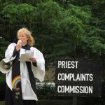 female priest with loud hailer standing next to a sign that says 'Priest complaints commission'