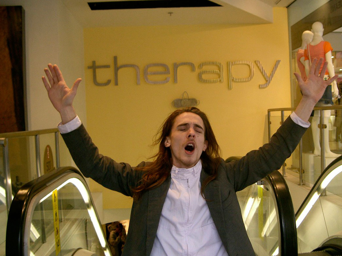 a white man with long hair at the top of an escalator with his arms raised