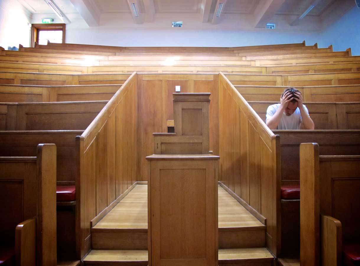 looking out from a lectern in a lecture hall a man sits on the right with his head in his hands