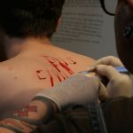 the naked back of a white man with blood on it as another man with a tattooed arm carves words into it with a scalpel
