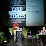man standing at a lectern and woman in a wheelchair at a table on stage with a projection behind reading "Crip Times"