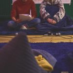 Two people sit cross legged next to each other on a fluffy blue rug. One is reading from a script.