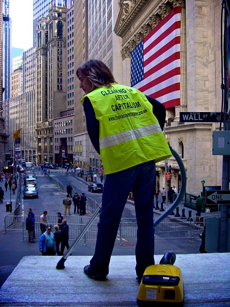 a man wearing a hi vis vest with "cleaning up after capitalism" printed on the back, is vacuuming in Wall Street