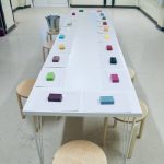 a long white table with different coloured and smelling soaps laid out on it