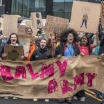 A group of young people hold a hessian sign with the words "Balmy Army" written on it in pink letters. They are marching as part of a busy protest, and a crowd is seen behind them holding handmade cardboard placards.