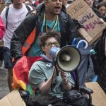 A young person in a wheelchair shouts into a megaphone as part of the Balmy Army March through Manchester. People hold handmade cardboard placards in the background.