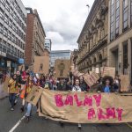 Young people are shown leading a march for mental health through the streets of Manchester, holding a banner that reads "Balmy Army."